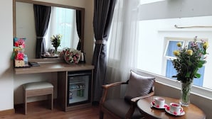 Habitación de hotel con un escritorio de madera, una silla, un refrigerador pequeño, una mesita de noche con tazas y un jarrón de flores.