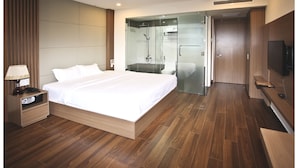 Una habitación de hotel moderna con una cama grande, una mesita de noche con lámpara, un televisor y una zona de ducha con mampara de vidrio.