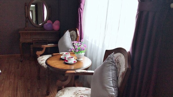 Una habitación con una mesa de comedor de madera, sillas con estampado floral, una pequeña mesita auxiliar con una tetera blanca y tazas, un espejo y cortinas moradas.