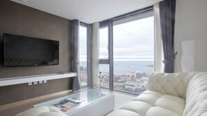A modern living room with a large window, a flat-screen TV, a white sofa, and a glass coffee table. - Eins Hotel (Seogwipo)