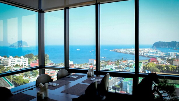 A dining area with a large window overlooking a coastal cityscape. - Eins Hotel (Seogwipo)