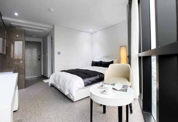 Una habitación de hotel moderna con una cama, una pequeña mesa redonda con una taza y una revista, una silla y un ventanal con vistas a la ciudad.