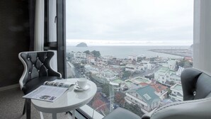 A room with a view of a cityscape and the sea. There is a table with a magazine and a cup. - Eins Hotel (Seogwipo)