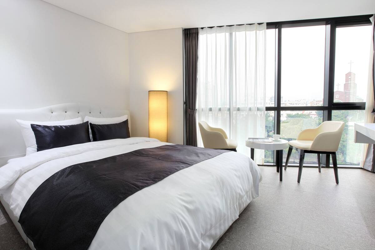 Une chambre d’hôtel avec un grand lit, une table de chevet, une chaise et une vue sur la ville grâce à de grandes fenêtres.
