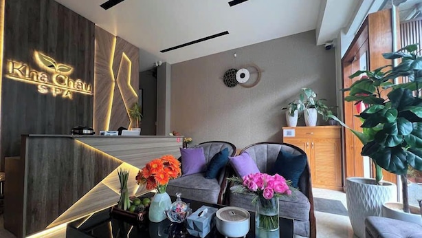 Een receptie met een houten receptiebalie, een bank met kussens, een bijzettafeltje met decoratieve objecten en een grote potplant.