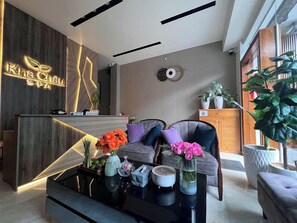 A reception area with a wooden reception desk, a sofa with cushions, a coffee table with decorative items, and a large potted plant. - Nam Xuan Premium Hotel (Xuan Huong - Da Lat)
