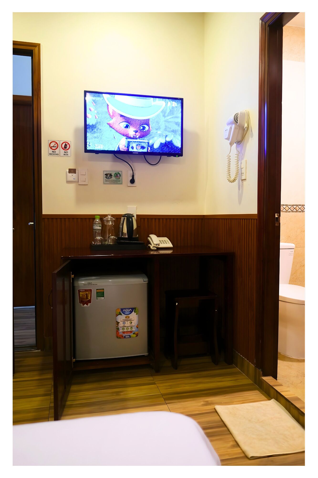 A hotel room with a television mounted on the wall, a small refrigerator, a desk with a telephone, and a door leading to the bathroom.