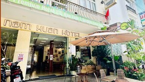 Een hotelentree met een bord waarop 'Nam Xuan Hotel' staat, terras met parasols en een motorfiets geparkeerd voor de deur.