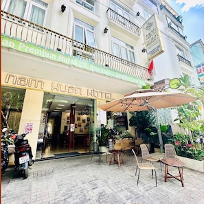 A hotel entrance with a sign reading 'Nam Xuan Hotel,' outdoor seating with umbrellas, and a motorcycle parked outside. - Nam Xuan Premium Hotel (Da Lat)