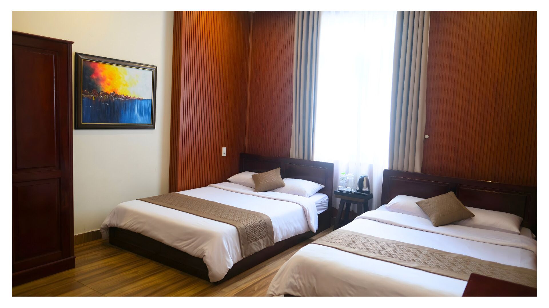 A hotel room with two beds, wooden paneling, a painting on the wall, and a window with curtains.