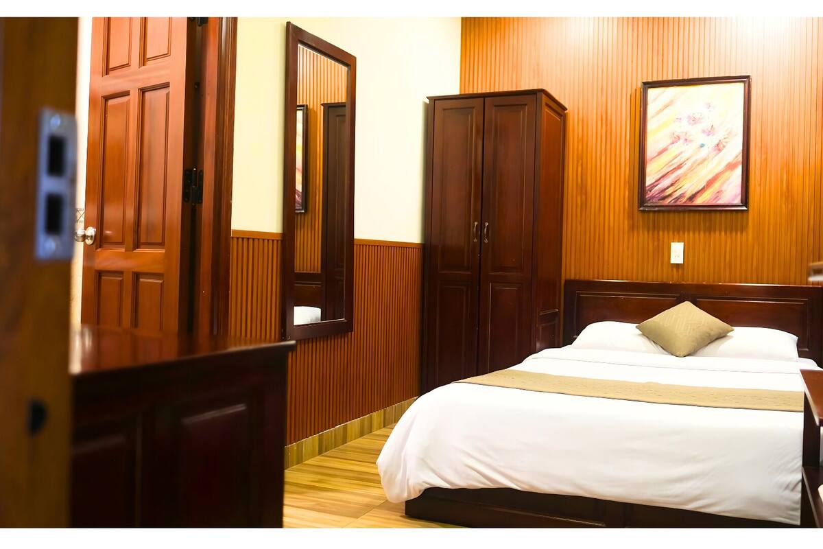A hotel room with a bed, wooden wardrobe, and a painting on the wall.