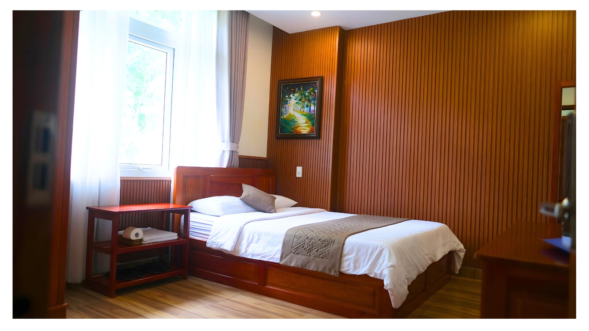 A bedroom with a wooden headboard, a bed with white bedding, a nightstand, a shelf with towels and slippers, a painting on the wall, and a window with curtains.