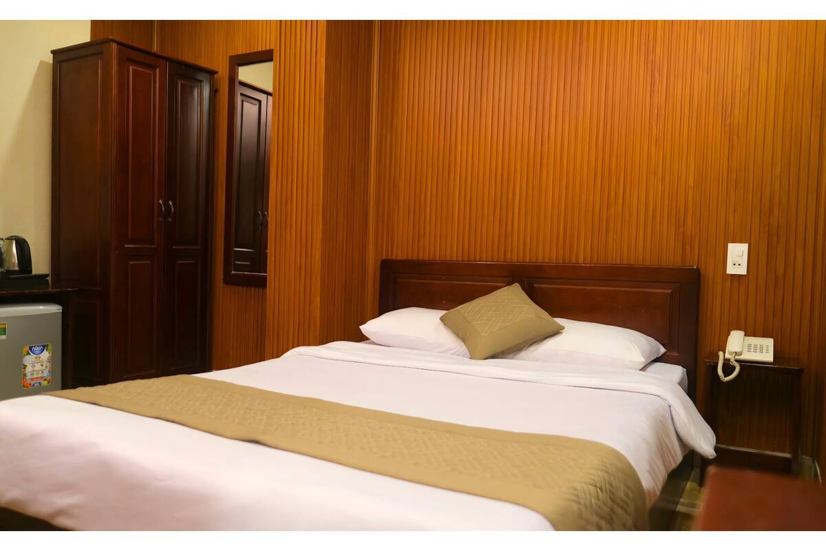 A hotel room with a bed, wooden paneling, a desk with a phone, and a refrigerator.