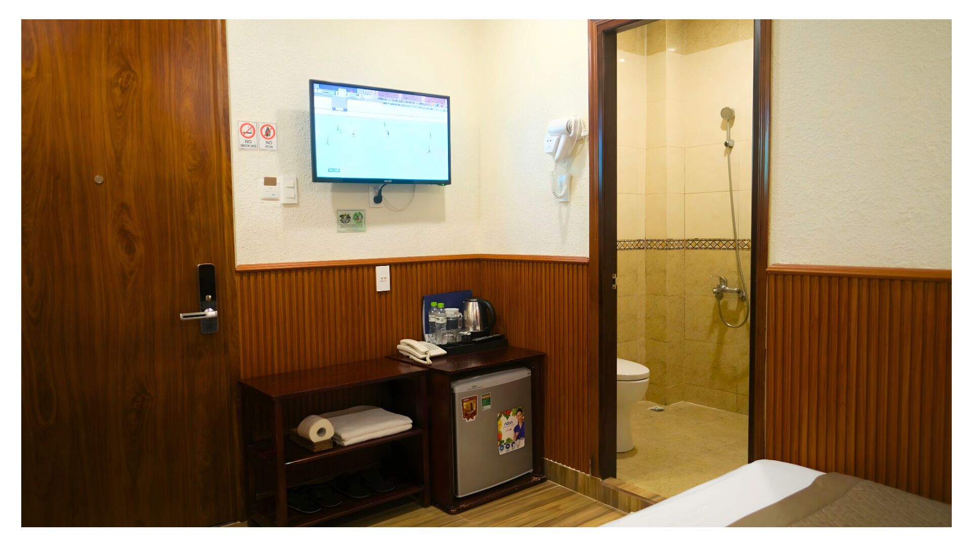 A hotel room with a wooden door, a desk with a phone and a small refrigerator, a toilet, a shower, and a television mounted on the wall.