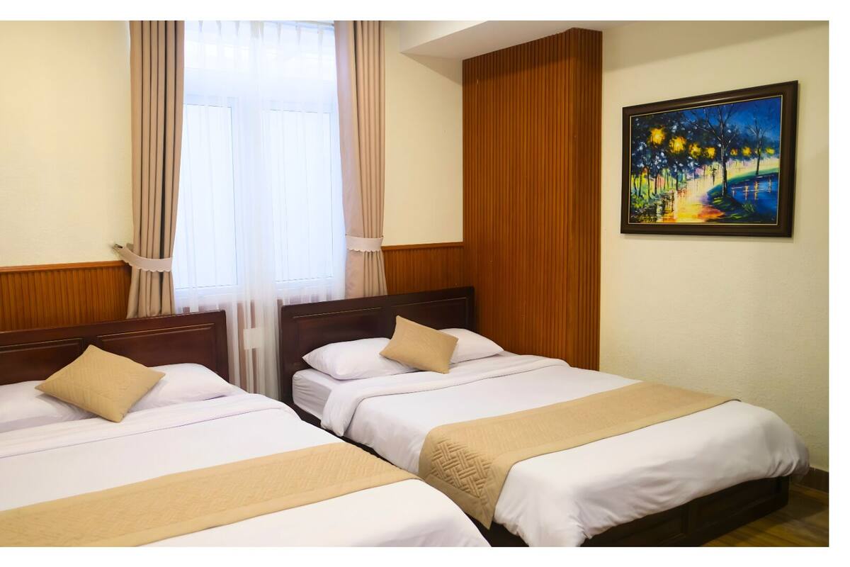 A hotel room with two beds, wooden paneling, a painting on the wall, and a window with curtains.