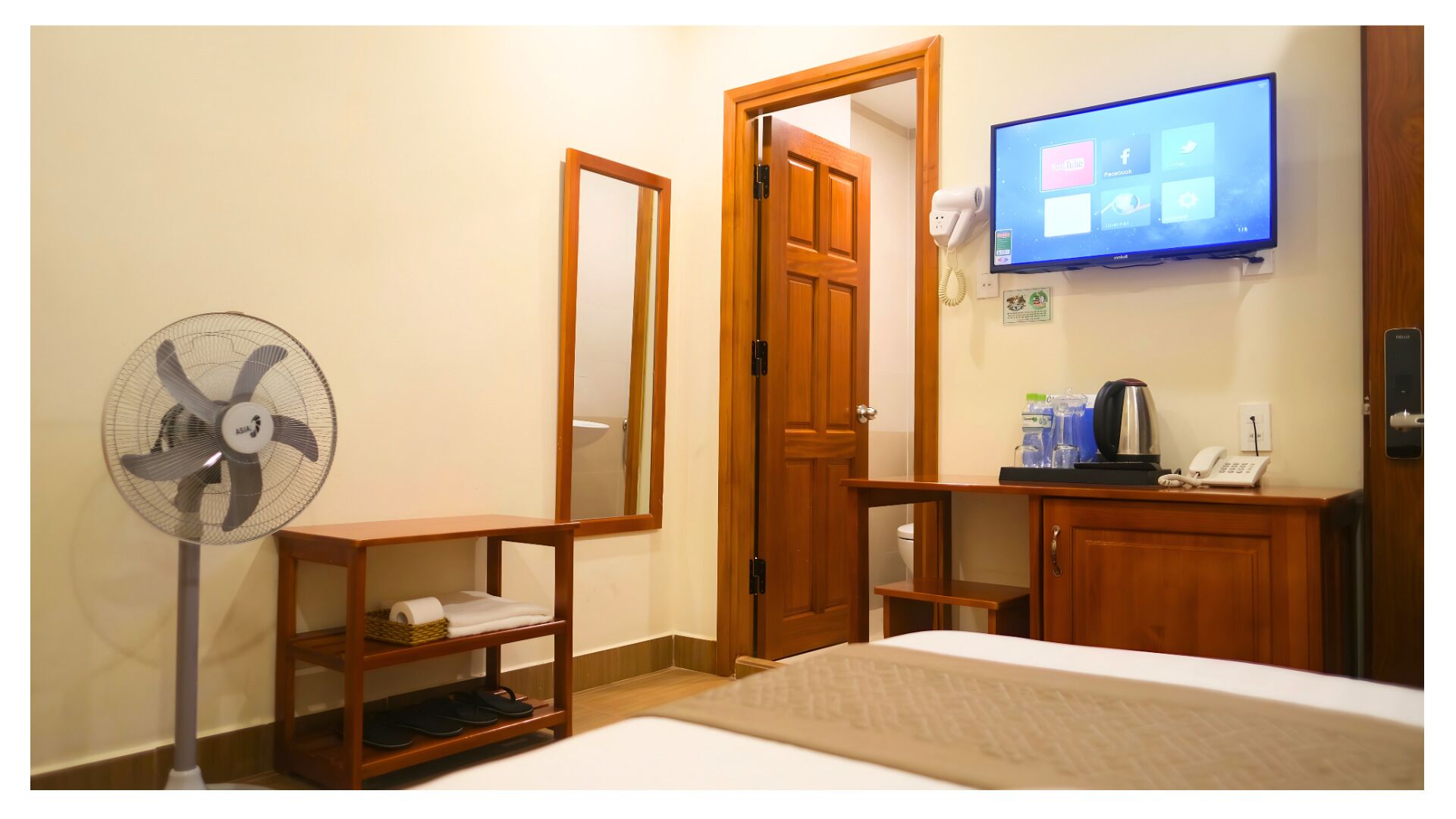 A hotel room with a bed, a wooden wardrobe, a desk with a telephone, a wall-mounted TV, a standing fan, and a door.
