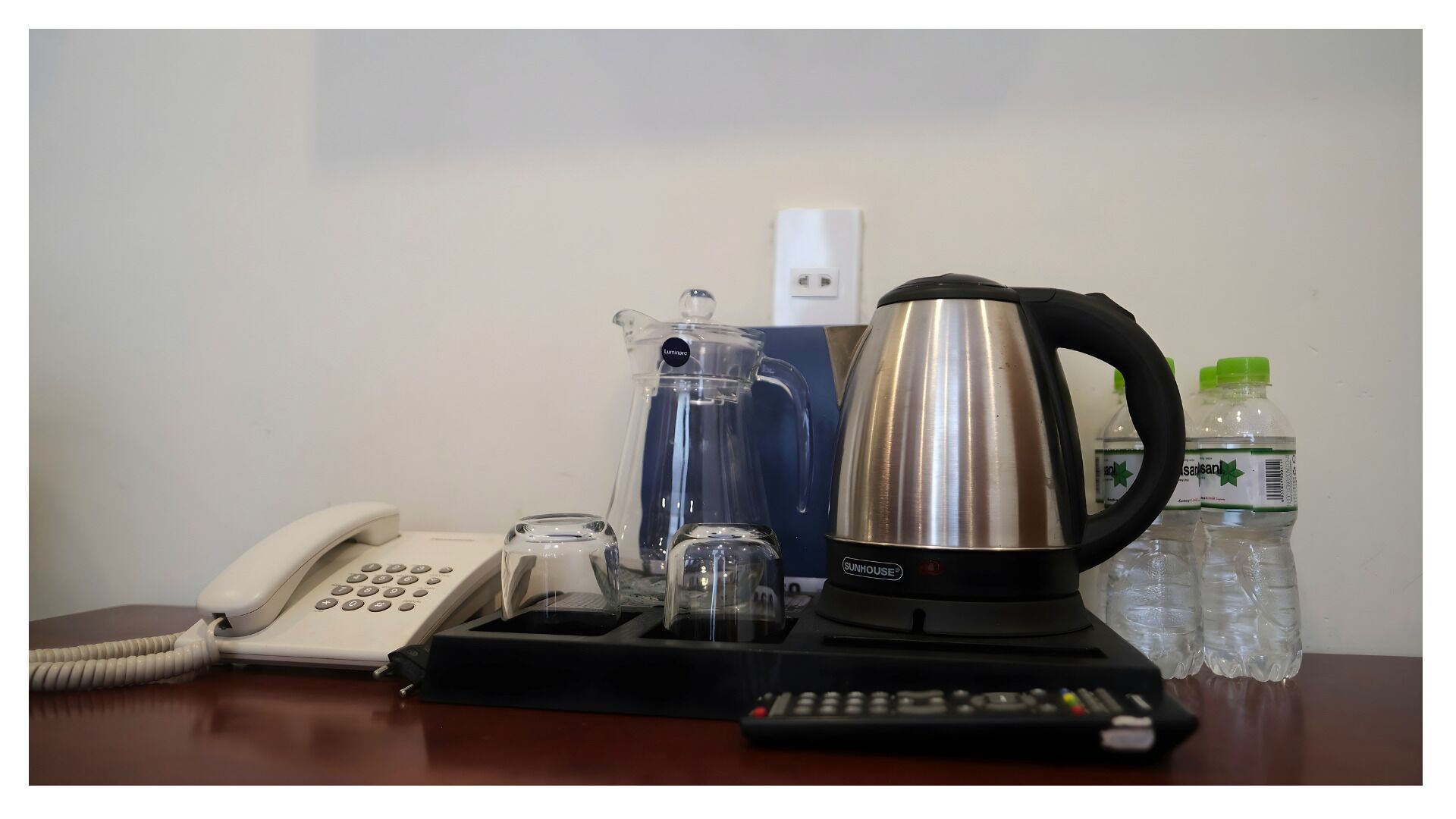 Une chambre d’hôtel équipée d’un téléphone, d’une carafe en verre, d’une bouilloire en acier inoxydable, d’une télécommande et de bouteilles d’eau.