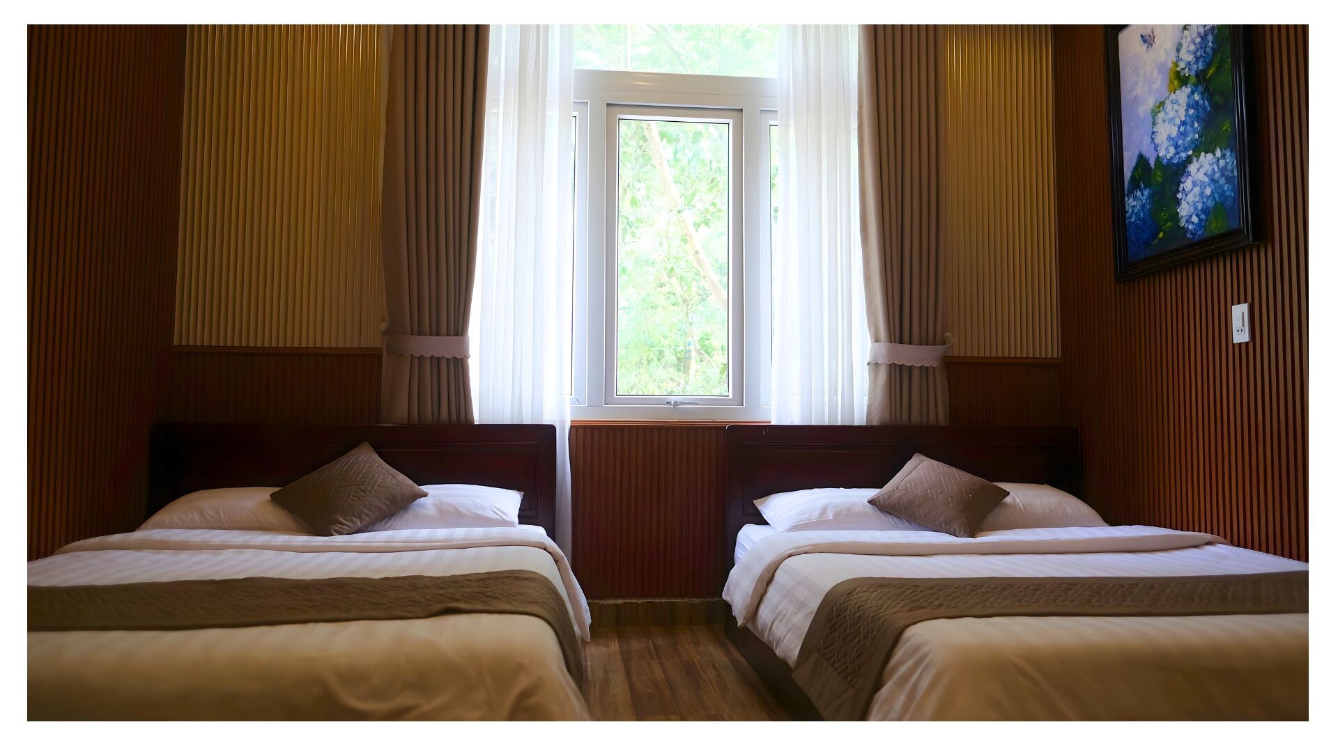 A room with two beds, a window with curtains, wooden paneling, and a painting on the wall.