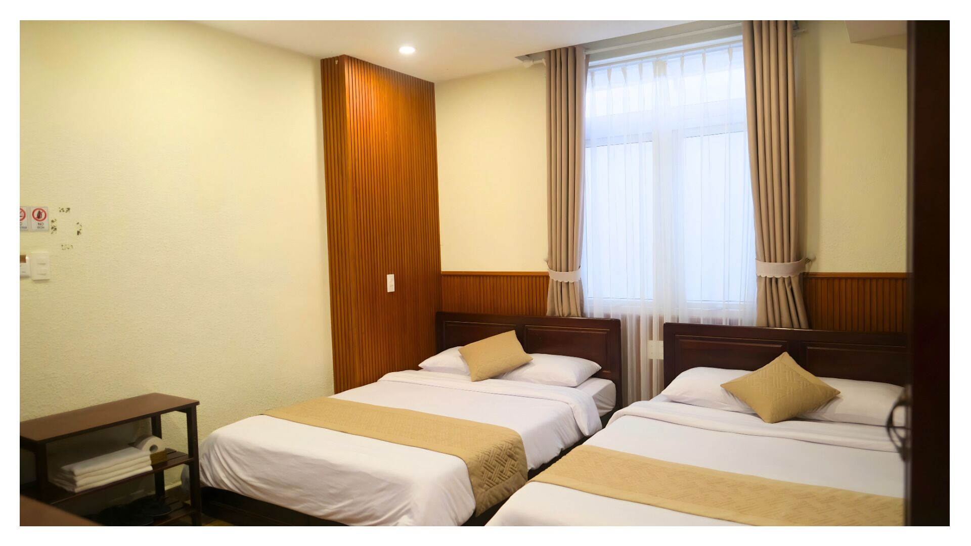 A hotel room with two beds, a wooden panel wall, a window with curtains, and a small table with a lamp.