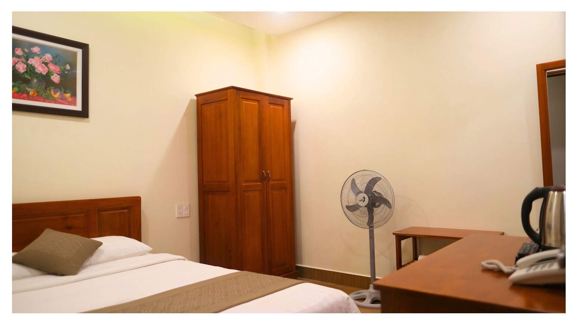 A hotel room with a bed, a wooden wardrobe, a standing fan, a desk with a telephone, and a framed picture on the wall.