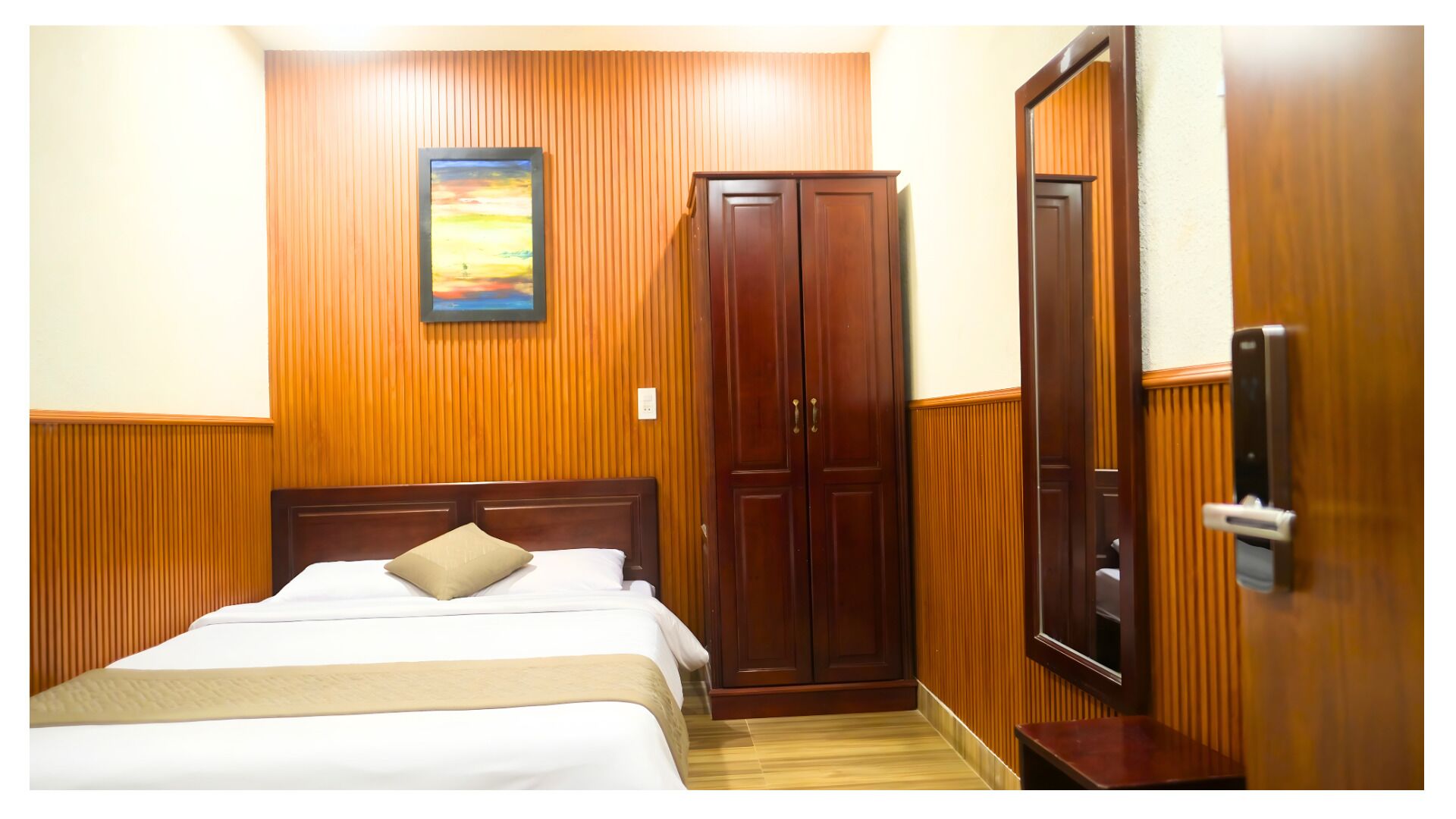 A hotel room with a wooden paneled wall, a bed with white linens, a wooden wardrobe, and a door with a handle.