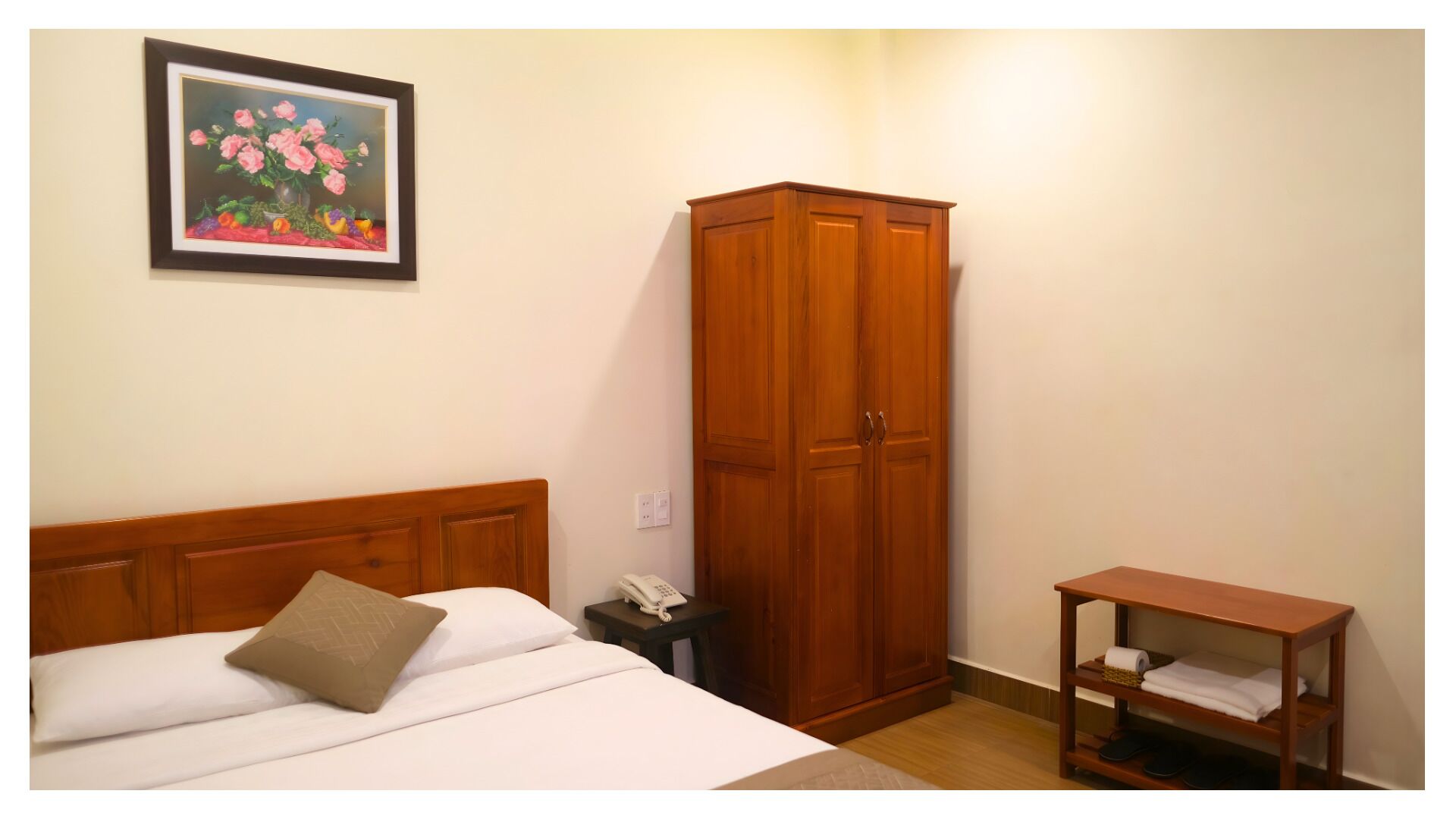 A bedroom with a bed, a wooden wardrobe, a bedside table with a phone, and a small wooden table with a towel and slippers.