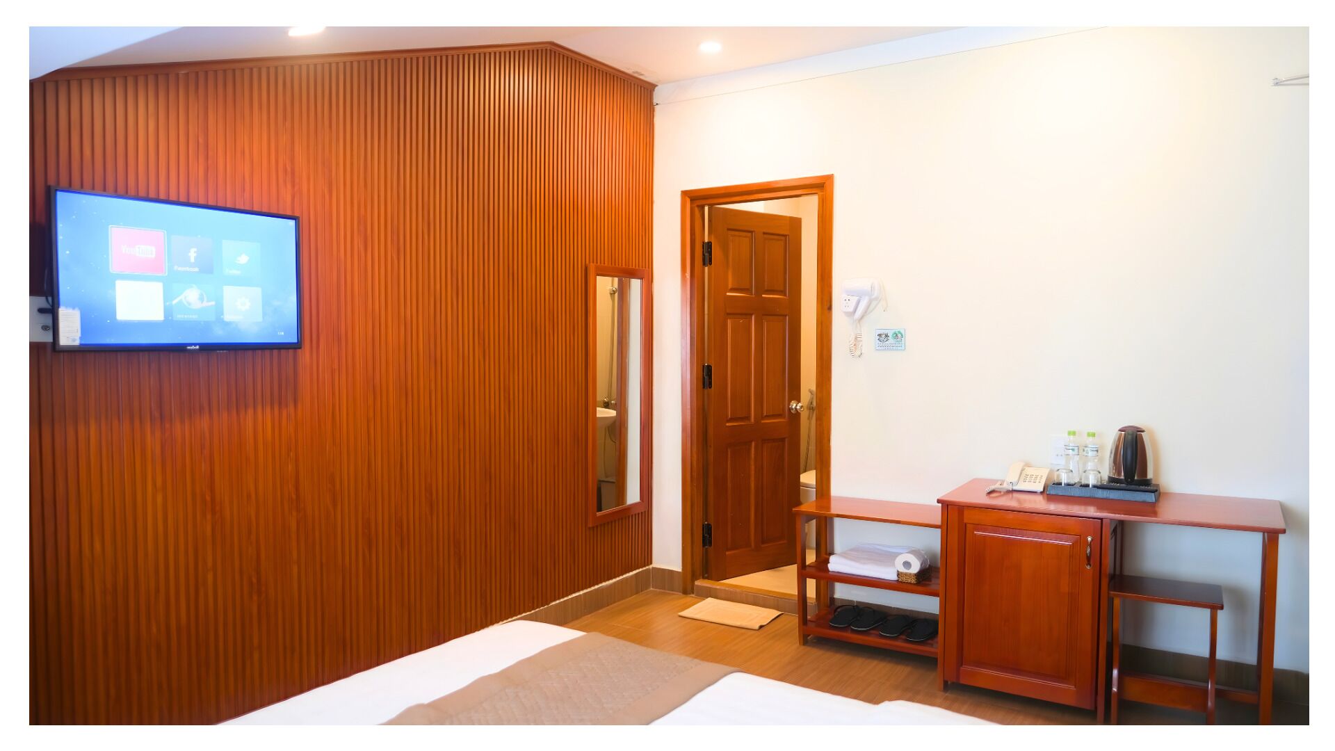 A hotel room with a wooden panel wall, a television mounted above a desk, a door leading to a bathroom, and a small table with a telephone and toiletries.