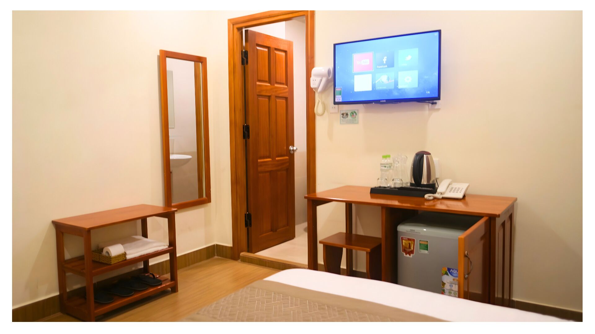 Habitación de hotel con un escritorio de madera, un refrigerador peque ño, un espejo, un televisor montado en la pared y una puerta.