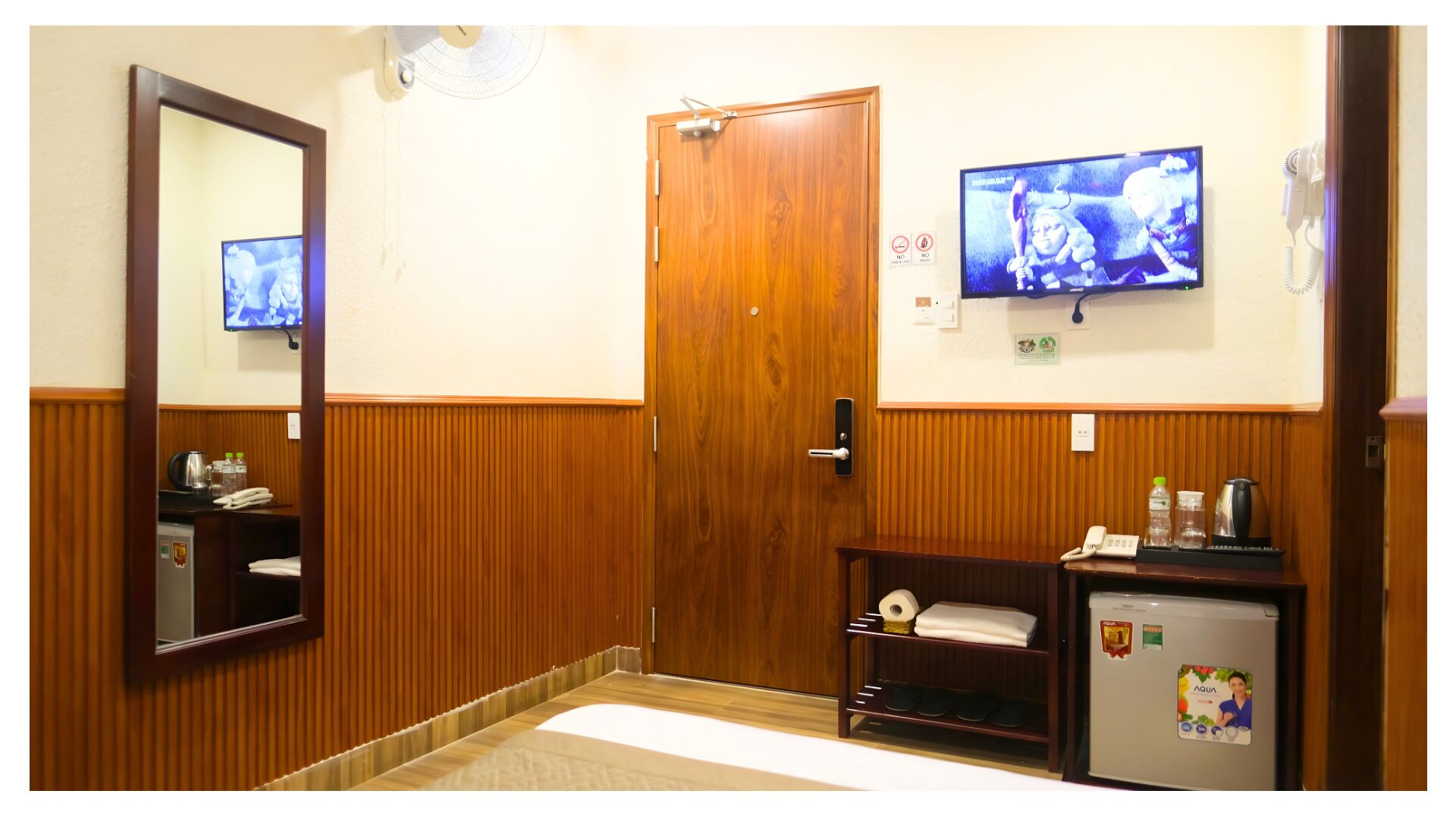 Un baño de hotel con puerta de madera, un espejo, televisor, lavamanos y refrigerador.