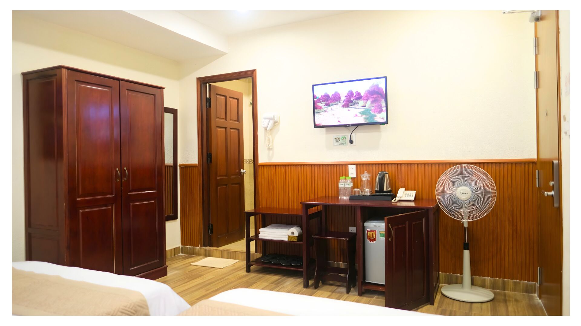 A hotel room with a bed, a desk with a computer, a TV, a fan, and a wardrobe.