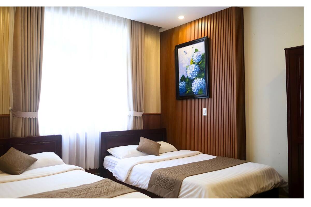 A hotel room with two beds, wooden paneling, a painting on the wall, and a window with curtains.
