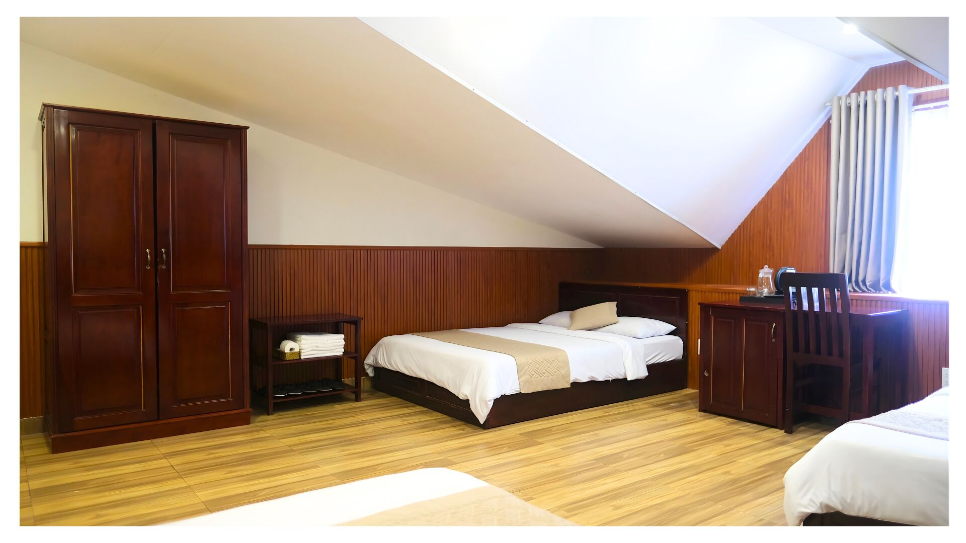 A bedroom with a sloped ceiling, wooden wardrobe, a bed with white bedding, a nightstand, and a desk with a chair.