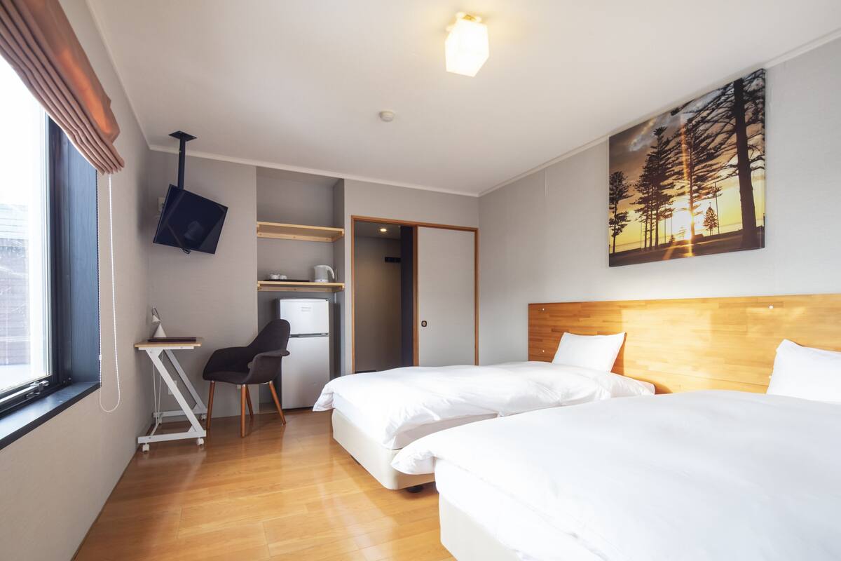 A hotel room with two beds, a desk, a chair, a TV, and a painting of trees.
