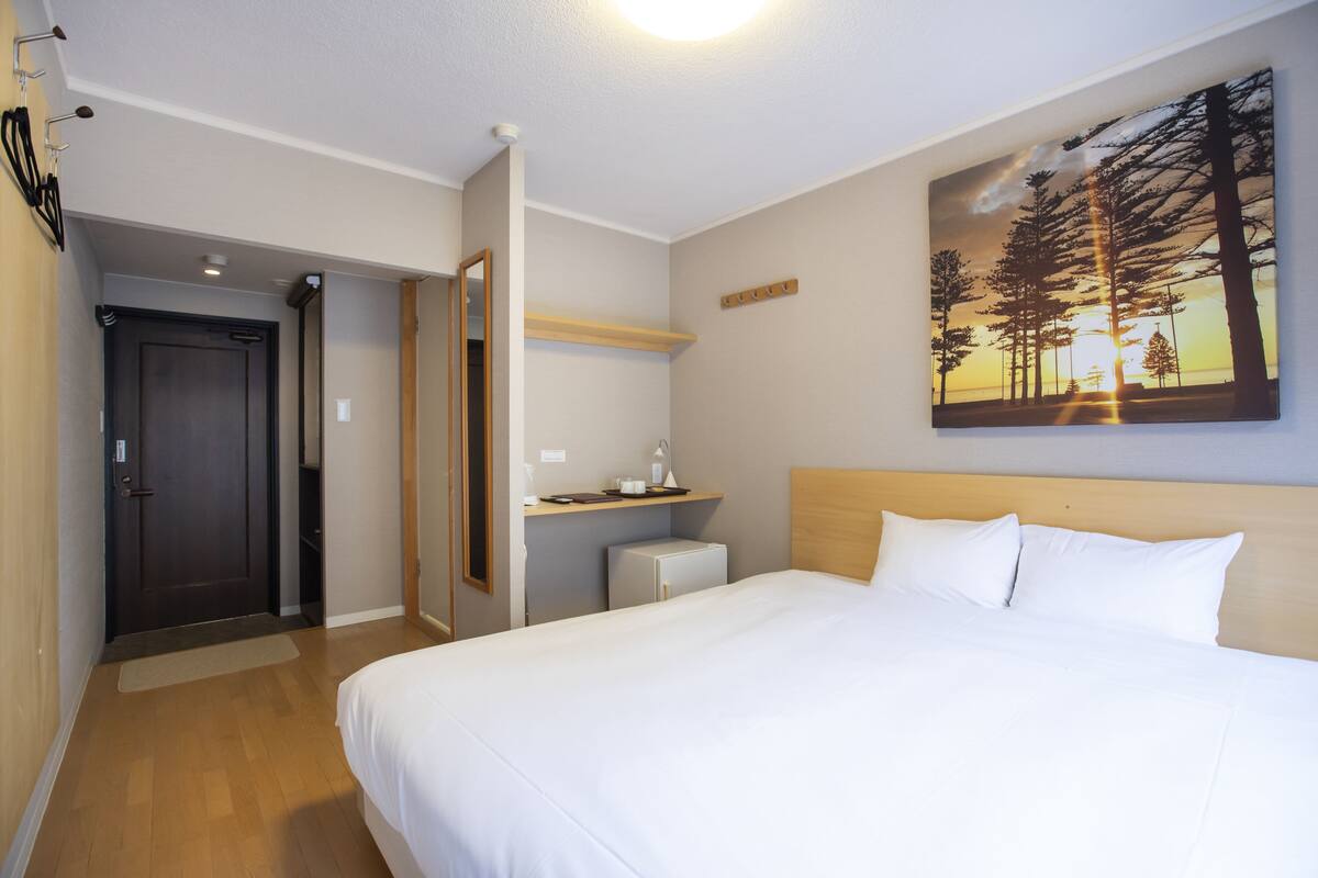 A hotel room with a bed, a desk, a chair, a nightstand, a mirror, a door, and a painting on the wall.