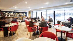 Un restaurante moderno con mesas redondas y sillas rojas, una zona de bar y grandes ventanales con vistas.