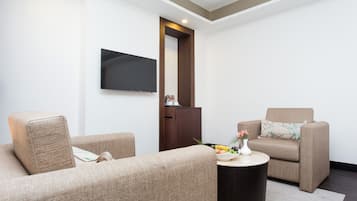 A modern living room with two beige sofas, a round coffee table, a flat-screen TV mounted on the wall, and a small side table with a vase and fruit.