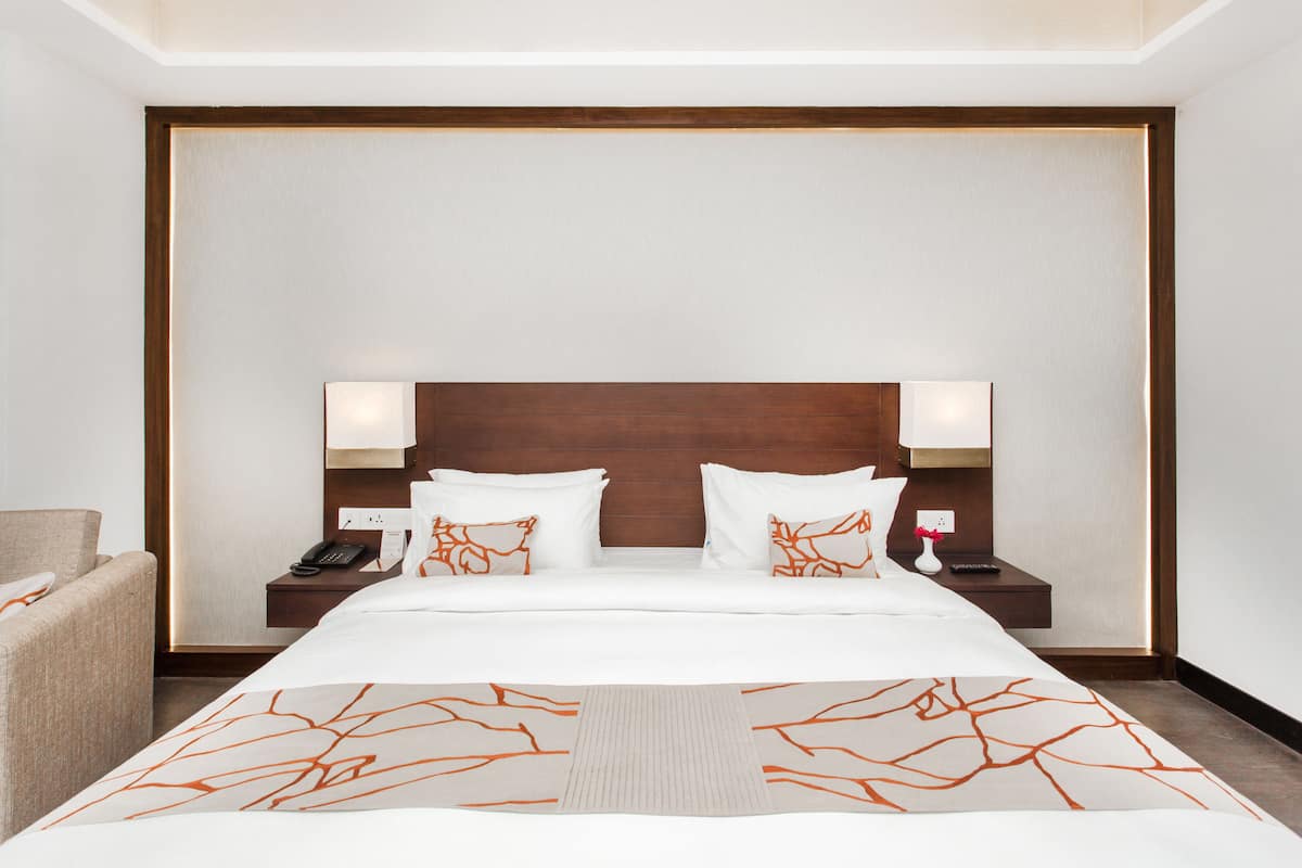 A hotel room with a large bed, two bedside tables, a nightstand, a chair, and a wall-mounted lamp.