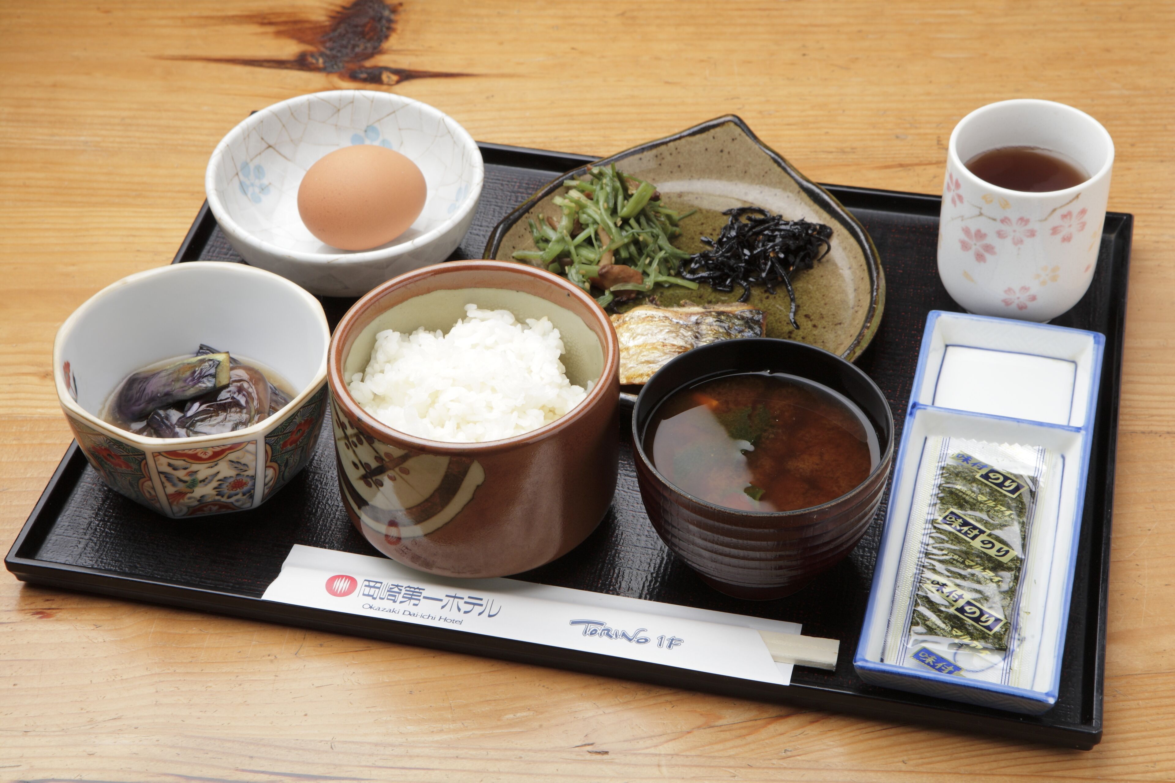 A tray with various Japanese dishes including rice, fish, and soup, along with a cup of tea and a small bowl of pickles.