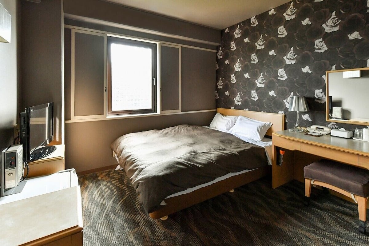 Ett hotellrum med en säng, ett skrivbord med en telefon, en spegel och ett fönster.