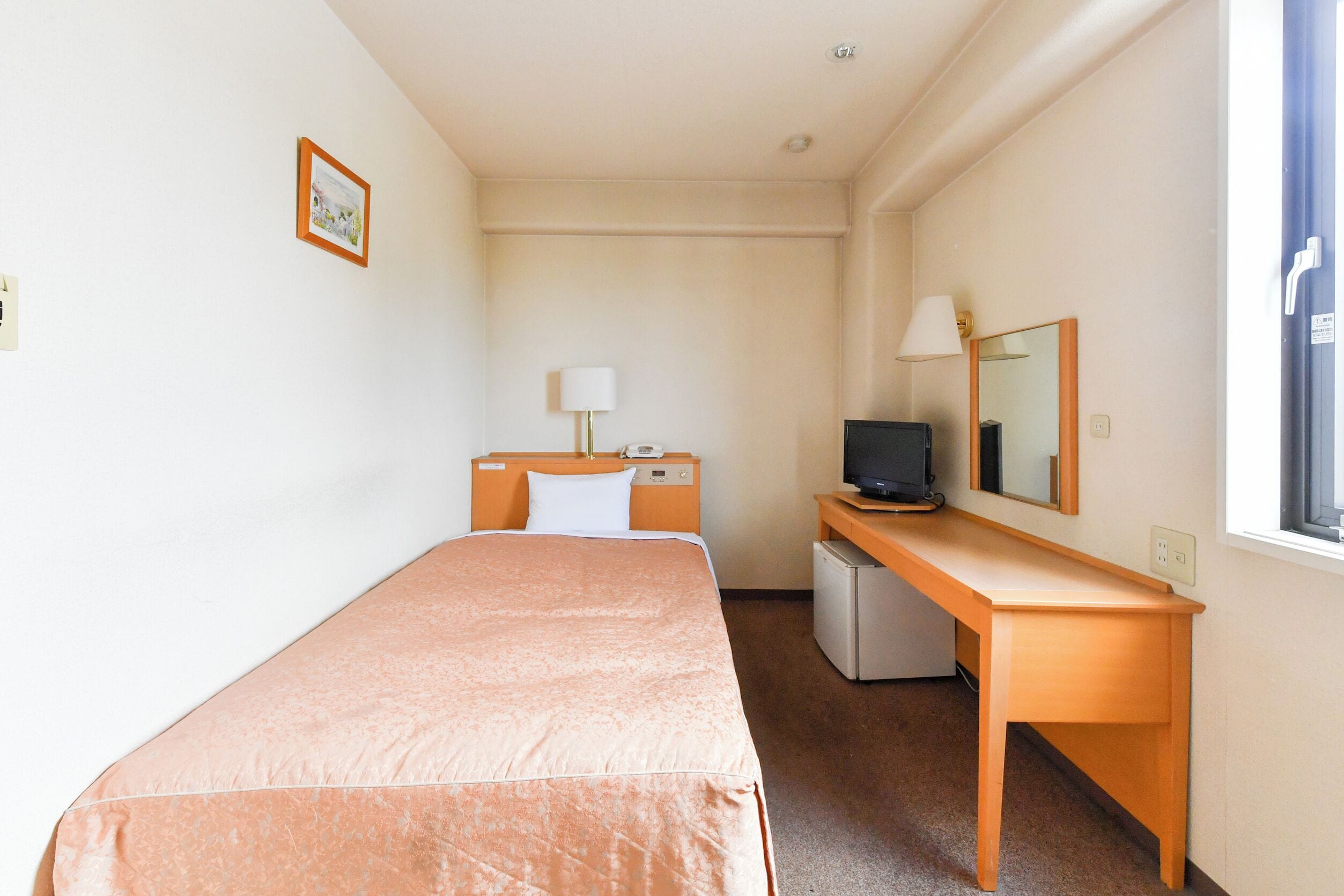 Une chambre d’hôtel avec un lit simple, un bureau avec un ordinateur, un petit réfrigérateur et une fenêtre avec des stores.