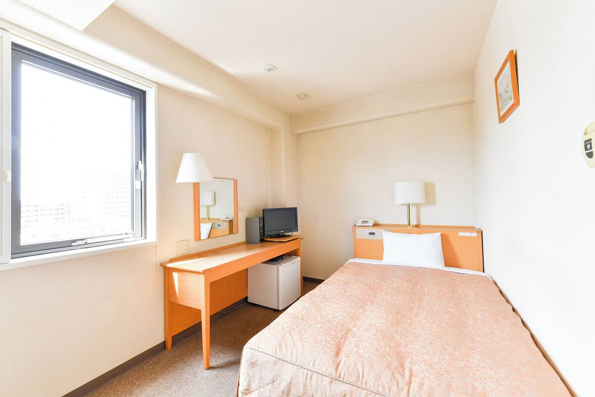 A hotel room with a bed, a desk with a computer, a small refrigerator, a window, and a wall-mounted lamp.
