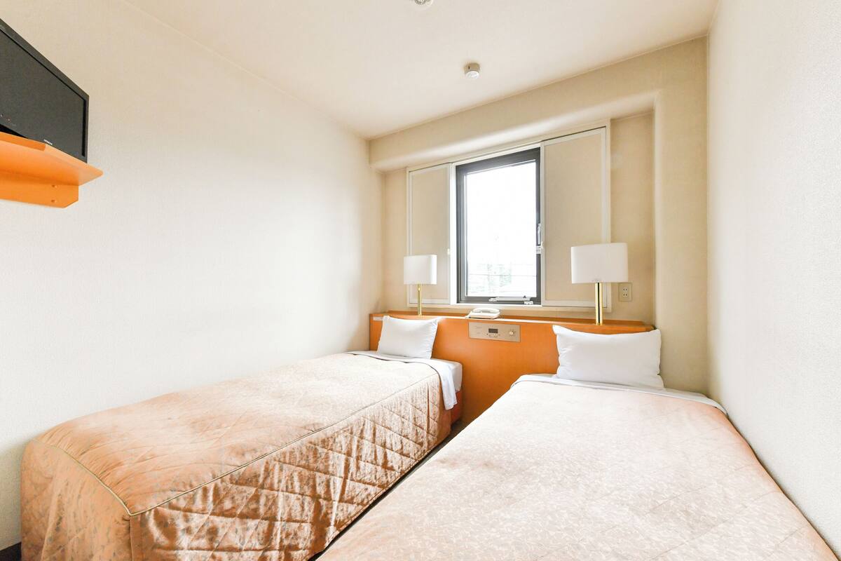 A hotel room with two single beds, a window with a view, a bedside table with a lamp, and a wall-mounted TV.