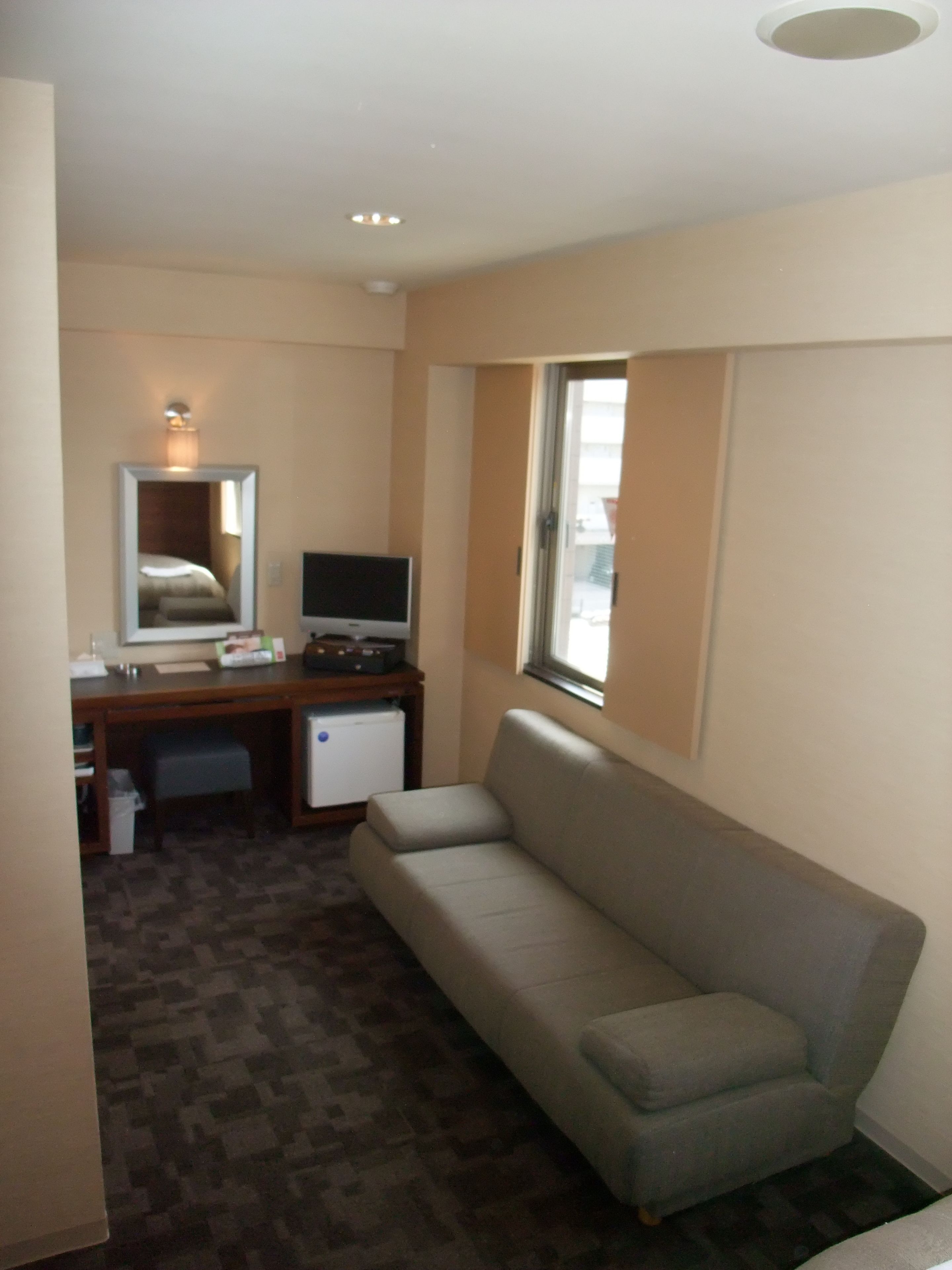 Habitación de hotel con sofá, escritorio con computadora, refrigerador pequeño, espejo y ventana.
