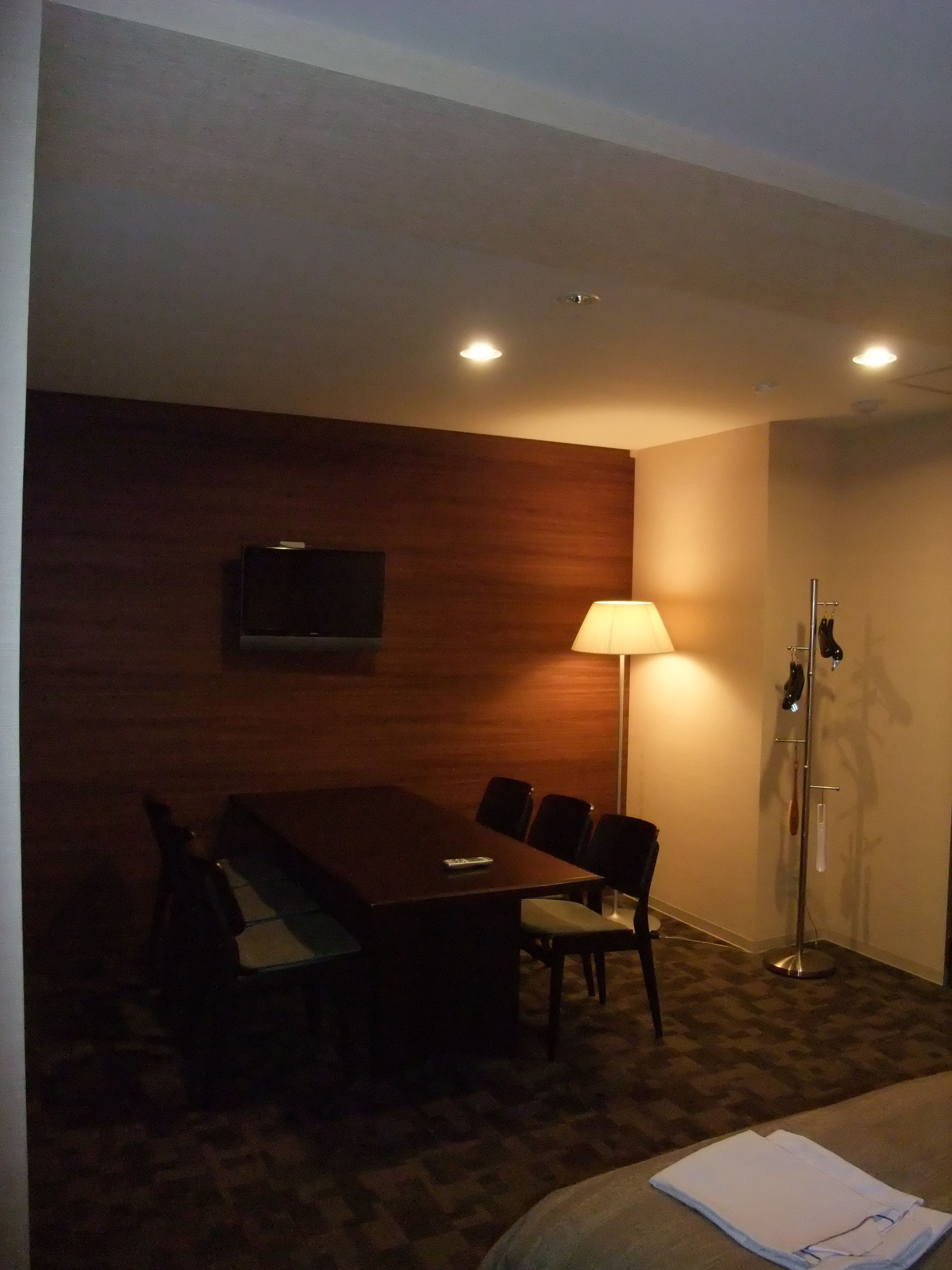 A conference room with a wooden table, chairs, a TV mounted on the wall, a standing lamp, and a coat rack with a mirror.