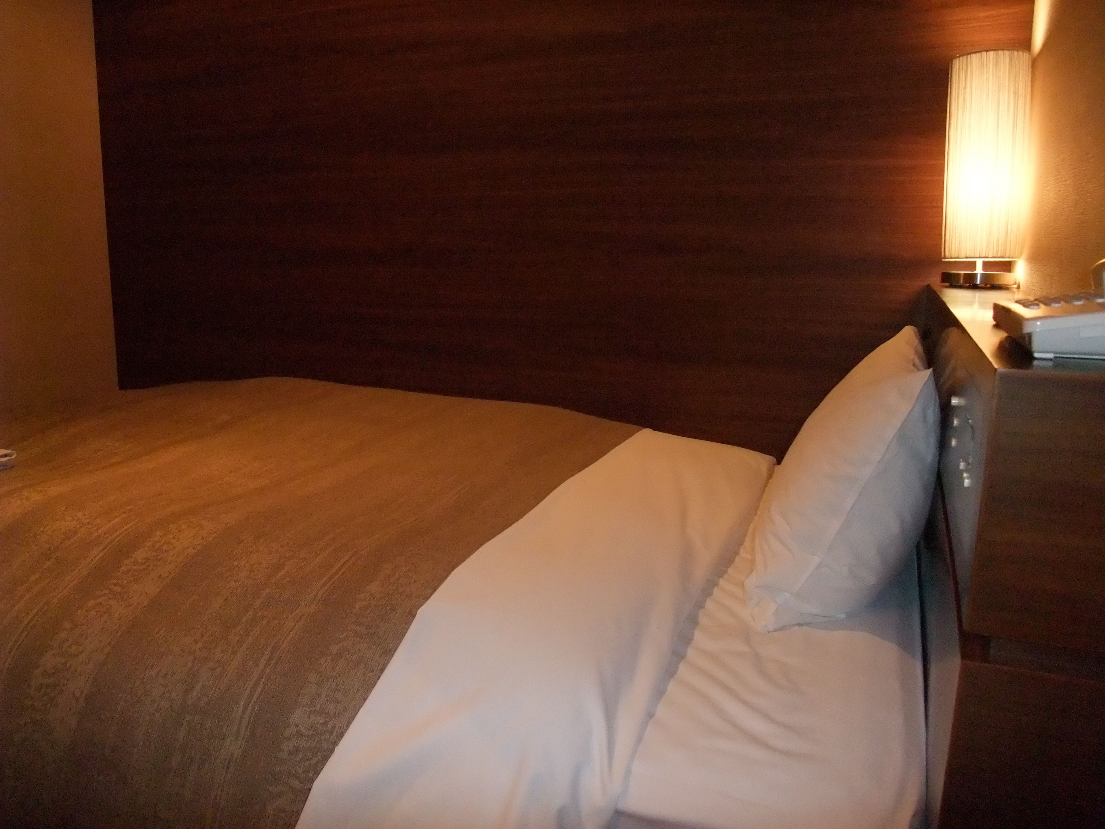 Ein Hotelzimmer mit einem hölzernen Kopfteil, einer weißen Bettdecke und einem Nachttisch mit Lampe.