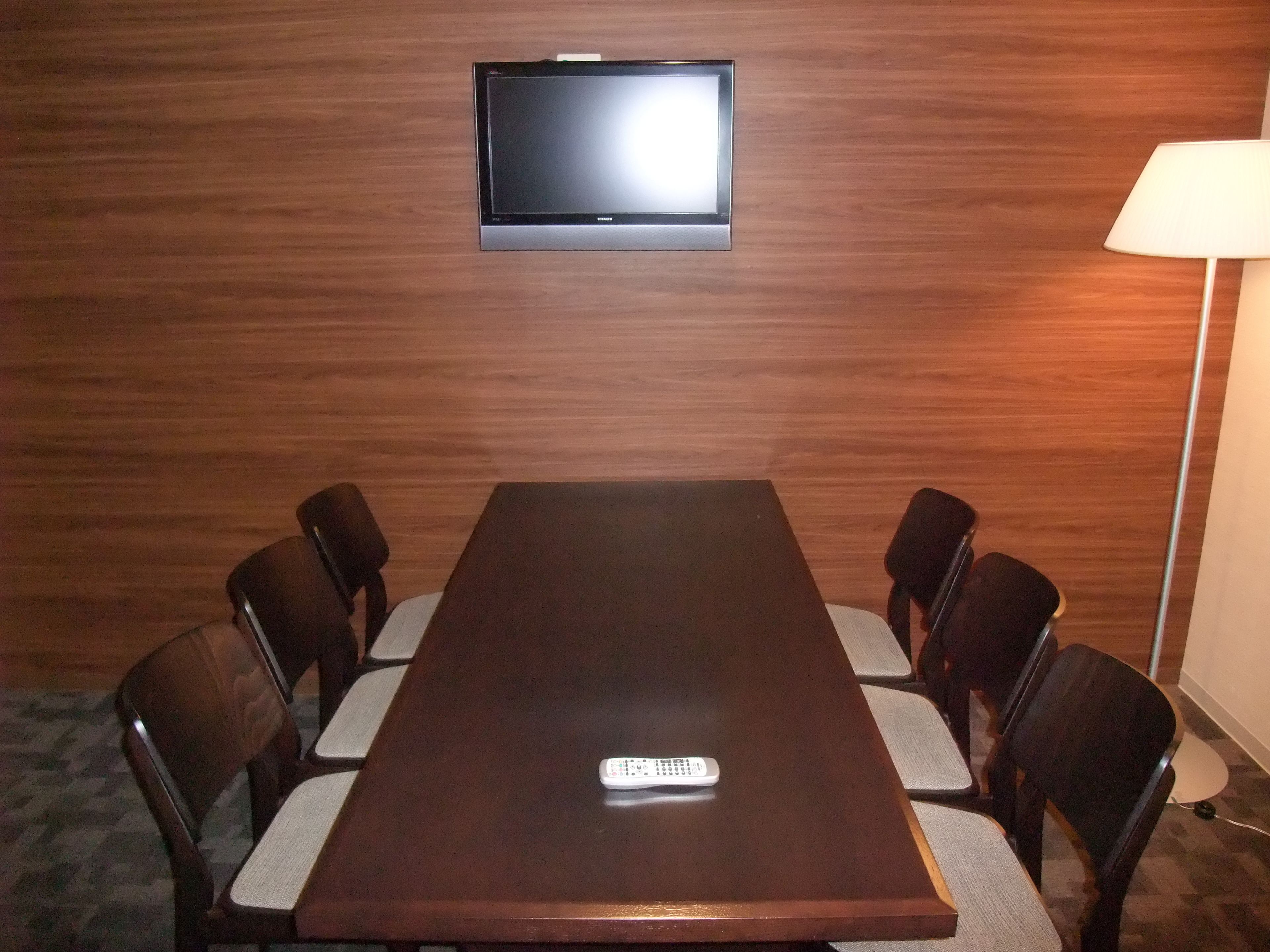 A conference room with a wooden table, six chairs, a television mounted on the wall, and a floor lamp.