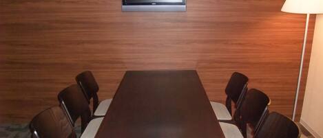 A conference room with a wooden table, six chairs, a television mounted on the wall, and a floor lamp.