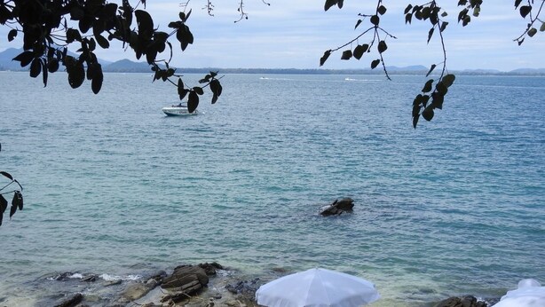 A coastal scene with rocky terrain, clear water, and a boat in the distance. There are tables with umbrellas, suggesting a dining area. - Sunrise Villas Seaview (Rayong)