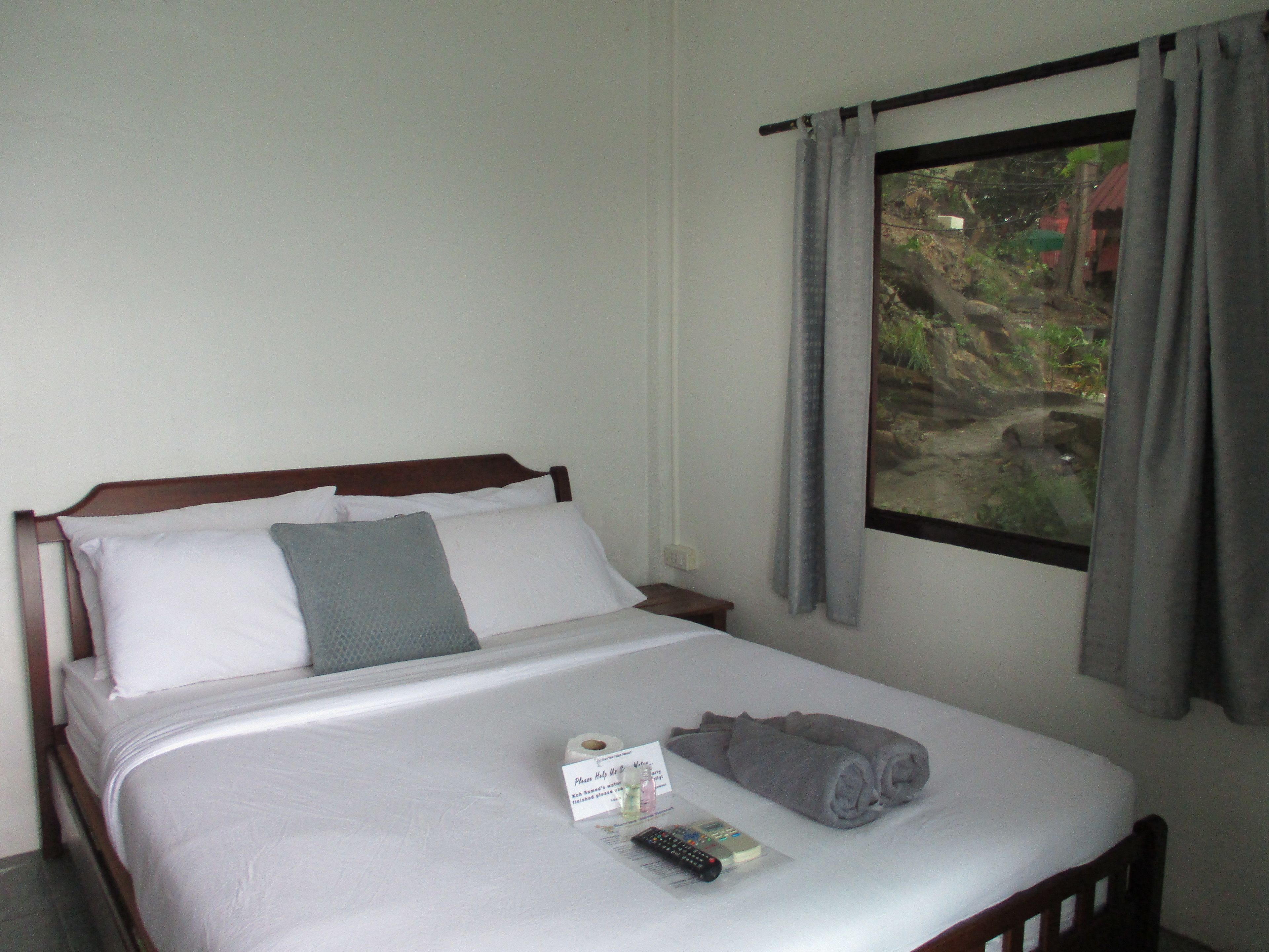 A neatly made bed with white linens, a grey pillow, and a rolled-up towel. A remote control and a card are placed on the bed.