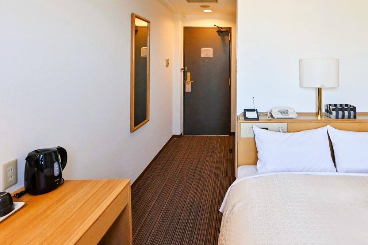 A hotel room with a bed, bedside table, lamp, and a view of the hallway.